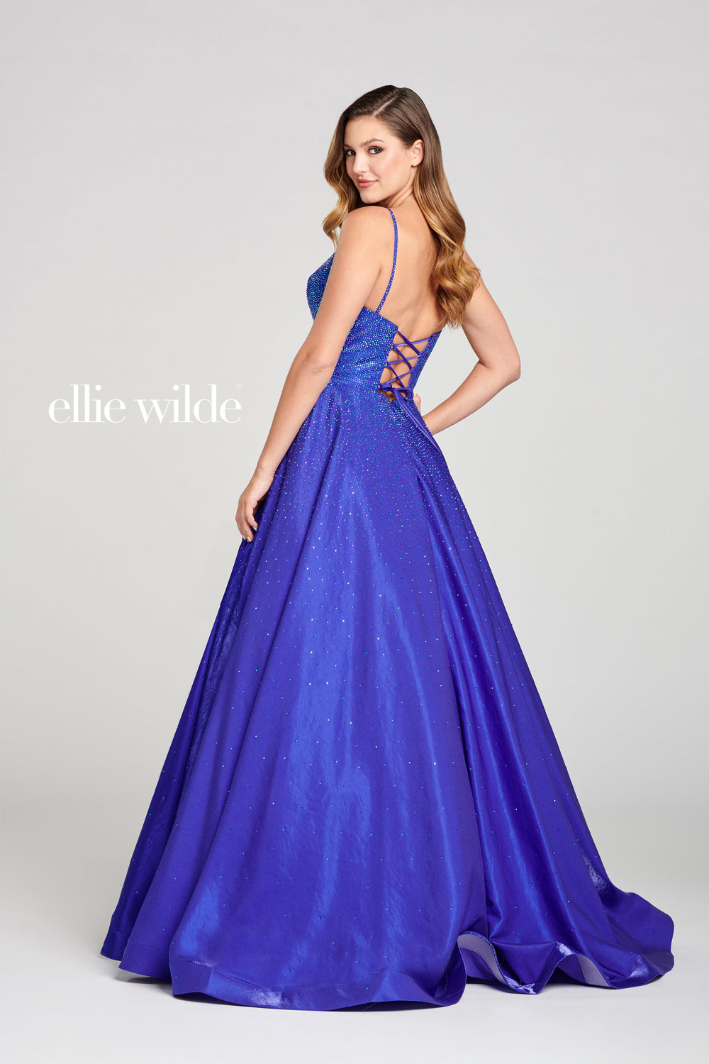 A woman models the Ellie Wilde Ew121005 Long Beaded Prom Ball Gown in royal blue, featuring a lace-up open back, beaded accents, and full A-line skirt against a light gray background. The